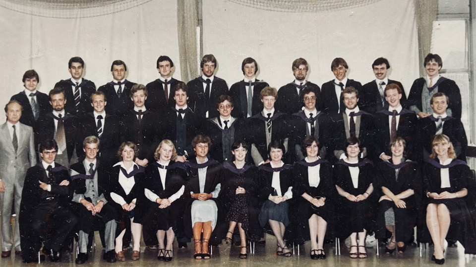 A group of people in academic gowns and formal attire pose in three rows for a photo against light-colored curtains.
